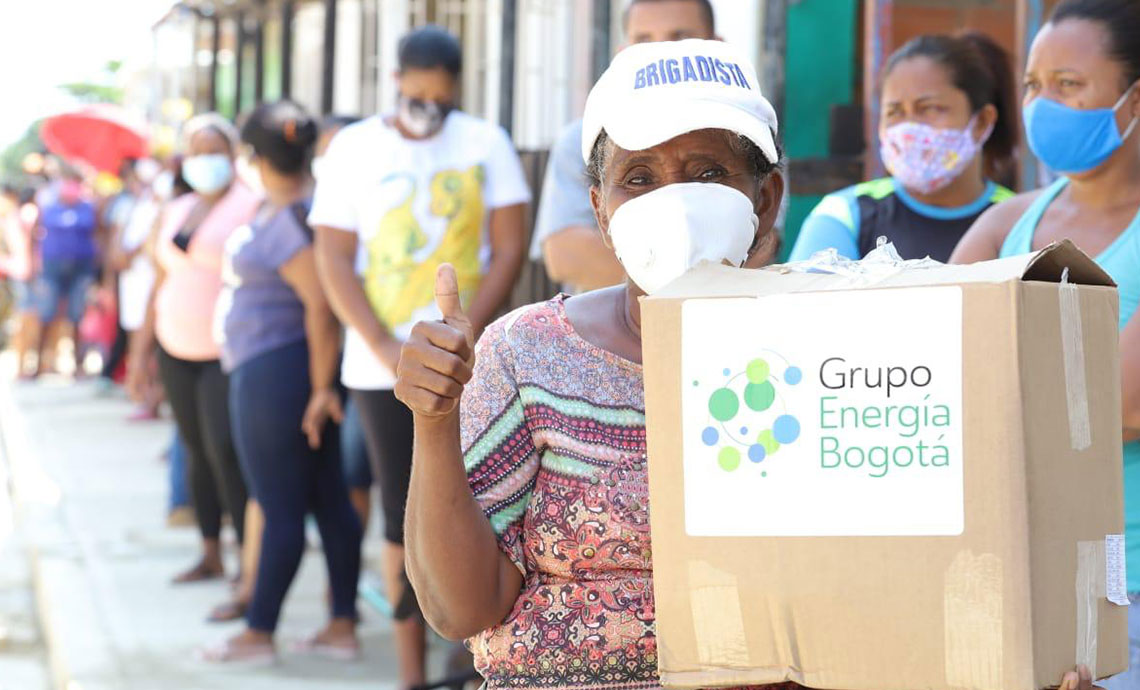 Woman with brigade member cap gives thumbs up and with the other hand carries a humanitarian aid package from GEB. Behind her, a line of people wait to receive their packages.