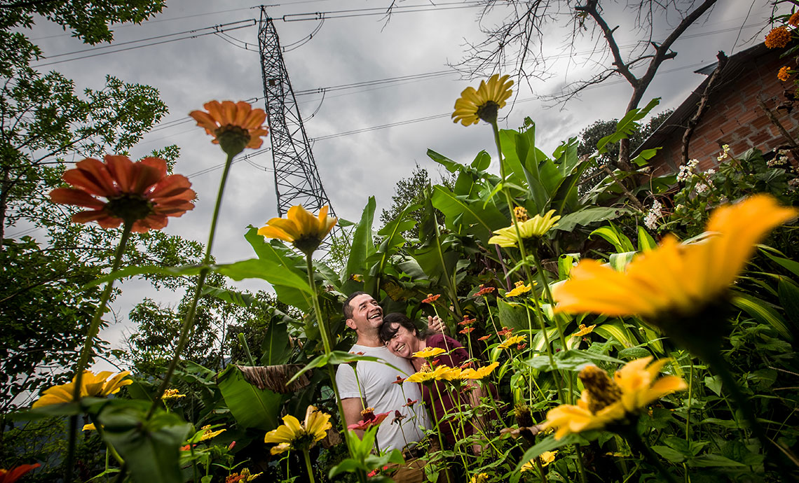 A couple embraces amidst yellow and orange flowers, with a GEB electricity tower in the background.