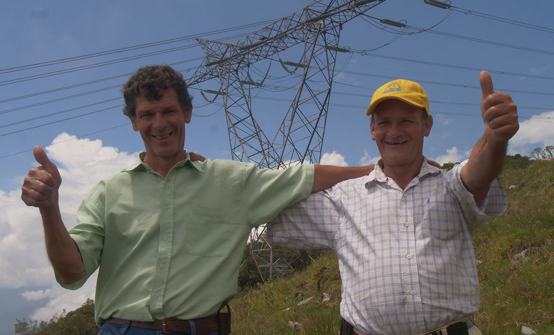 Dos hombres abrazados sonríen y tienen sus pulgares levantados, detrás de ellos hay una torre de energía de GEB.