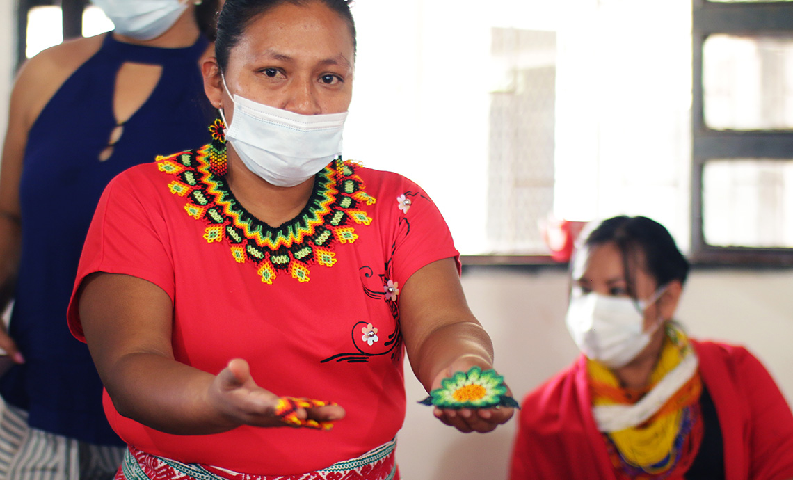 mujer indígena con tapabocas puesto y su ropa tradicional muestra con sus manos las artesanías que realiza.