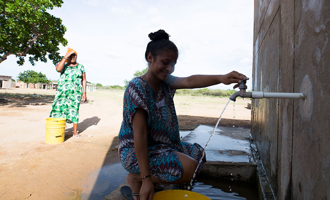 mujer joven está recogiendo agua, detrás de ella hay unas pocas casas en medio del desierto.