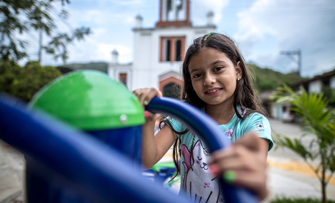  una niña se sonríe en el parque principal de un pueblo, detrás de ella está el atrio de la iglesia.