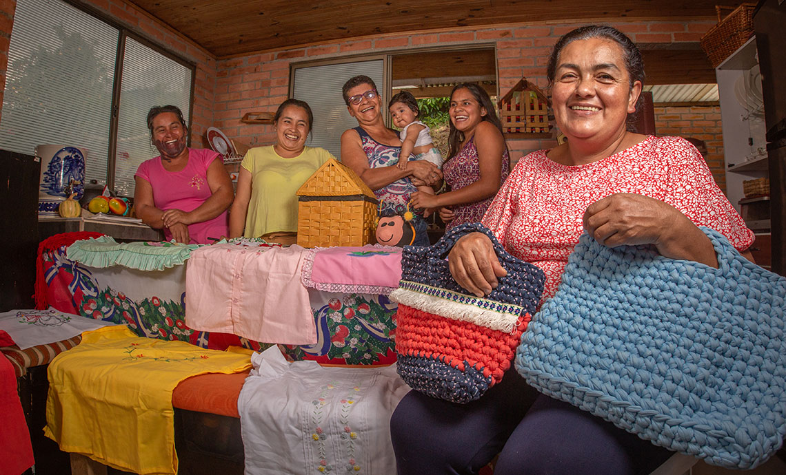Five smiling women from the Association of Women Victims of Violence at a house with handicraft knit work and one of them carrying a baby.