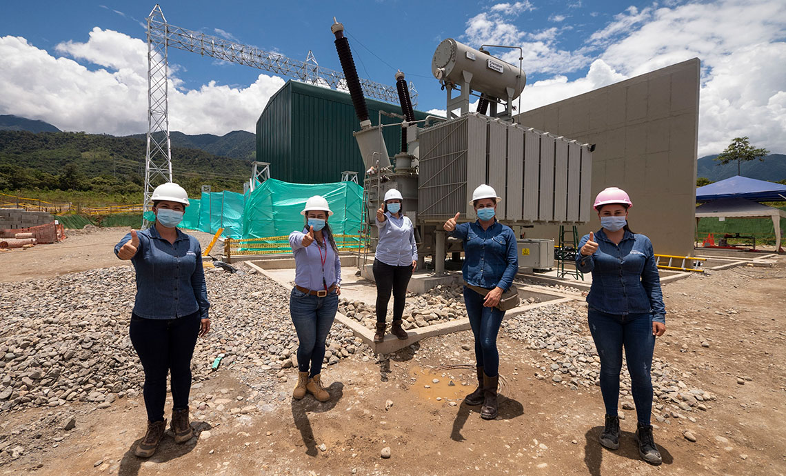 cinco mujeres, con sus uniformes y elementos de protección, están en la zona de la operación, detrás de ellas hay maquinaria, una planta y una torre de energía.