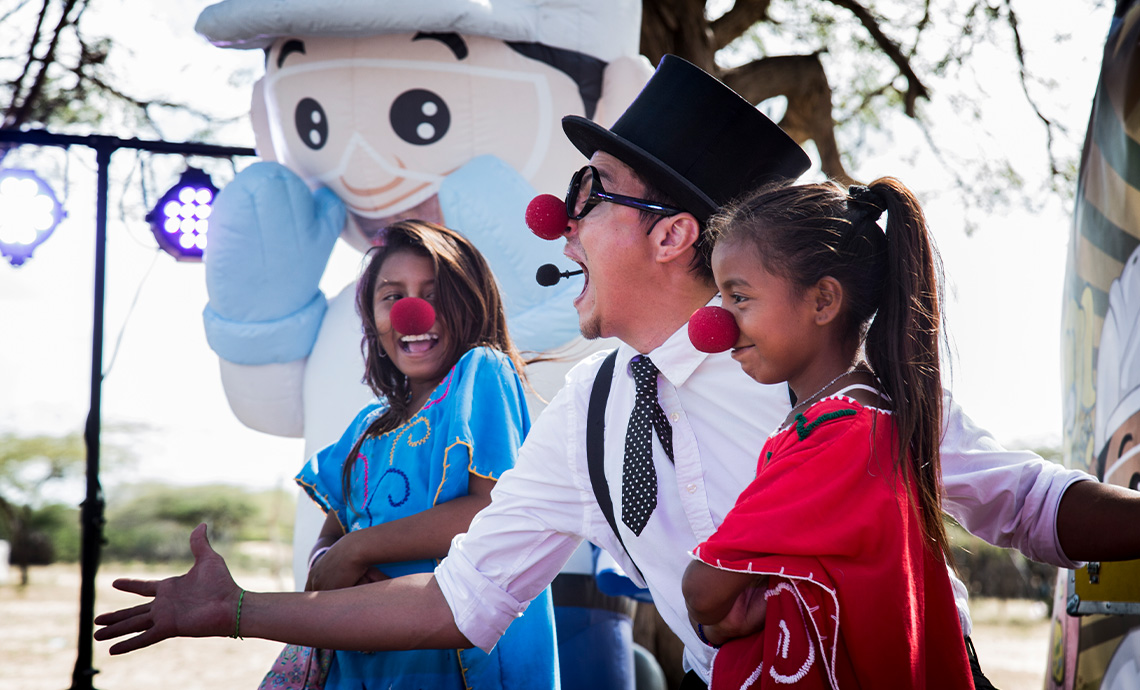 hombre de camisa blanca, corbatín negro con puntos blancos y sombrero negro con nariz de payaso hace reír a dos niñas wayús que también tienen puesta una nariz de payaso.